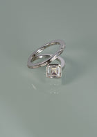 18k white gold bypass ring featuring a floating asscher cut diamond and tapering channel of baguettes band- against sea foam reflective background