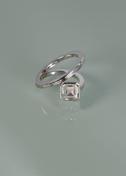 18k white gold bypass ring featuring a floating asscher cut diamond and tapering channel of baguettes band- against sea foam reflective background