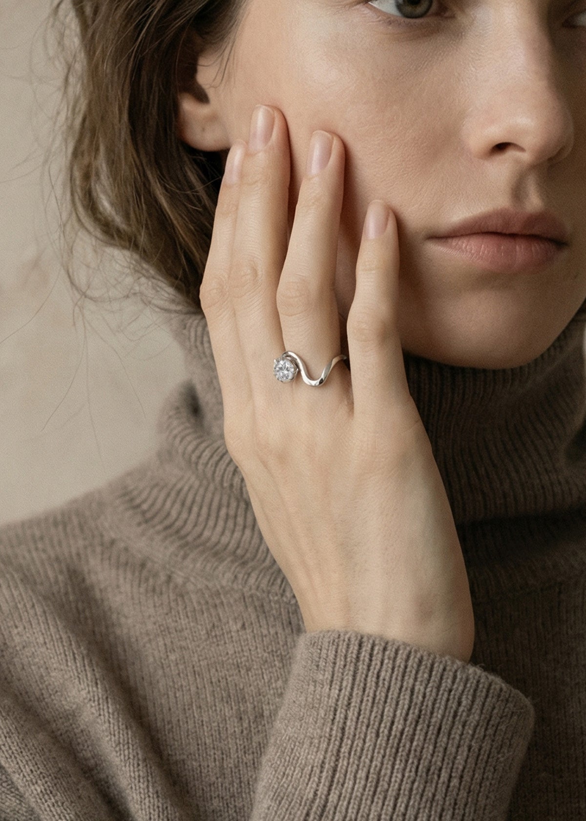 Model wearing a 18k white gold ring with a unique sculptural, flowing, wavy band nestling a large round cut diamond on her finger, wearing a brown sweater-KATKIM-engagement ring-fine jewelry