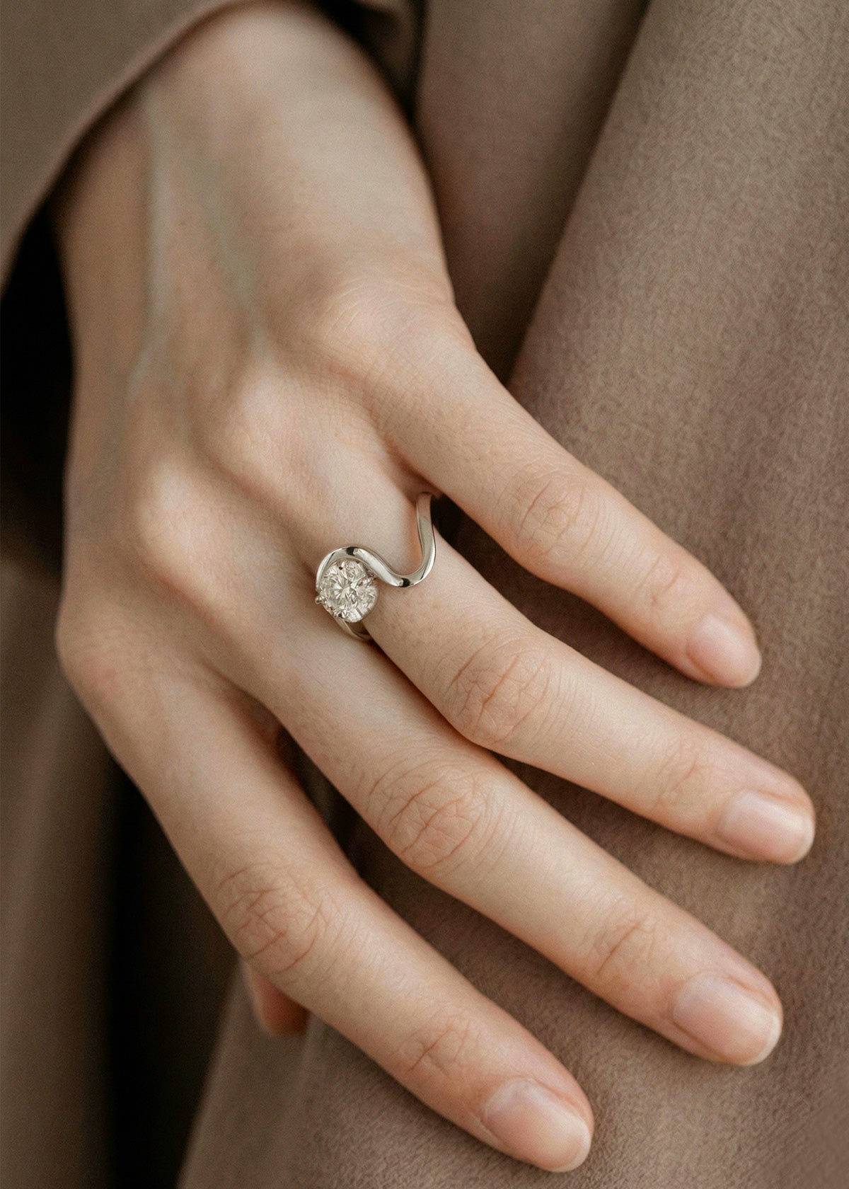 Close-up of a hand wearing a 18k white gold ring with a unique sculptural, flowing, wavy band nestling a large round cut diamond-hand is against a minimal, brown linen outwear-KATKIM-engagement ring-fine jewelry