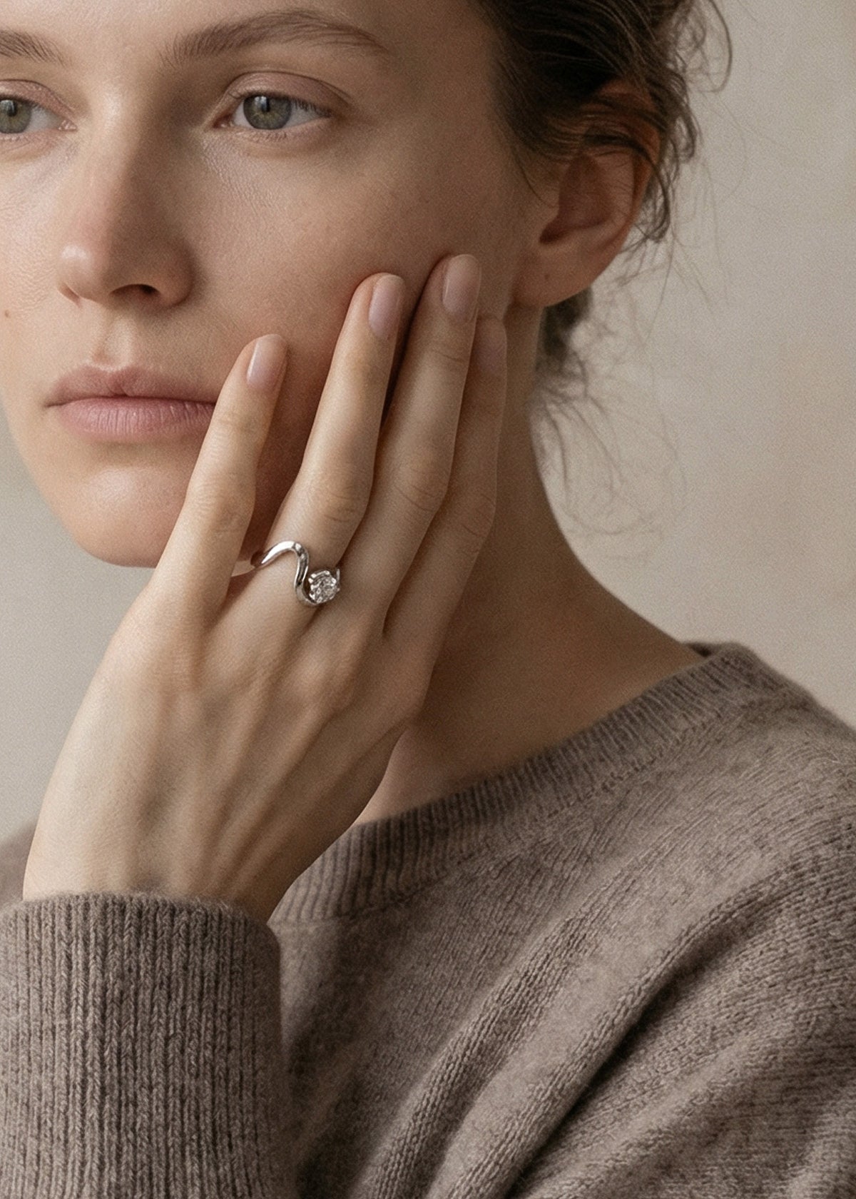 model wearing a 18k white gold ring with a unique sculptural, flowing, wavy band nestling a large round cut diamond-KATKIM-engagement ring-fine jewelry- holding her hand near her face against a neutral background