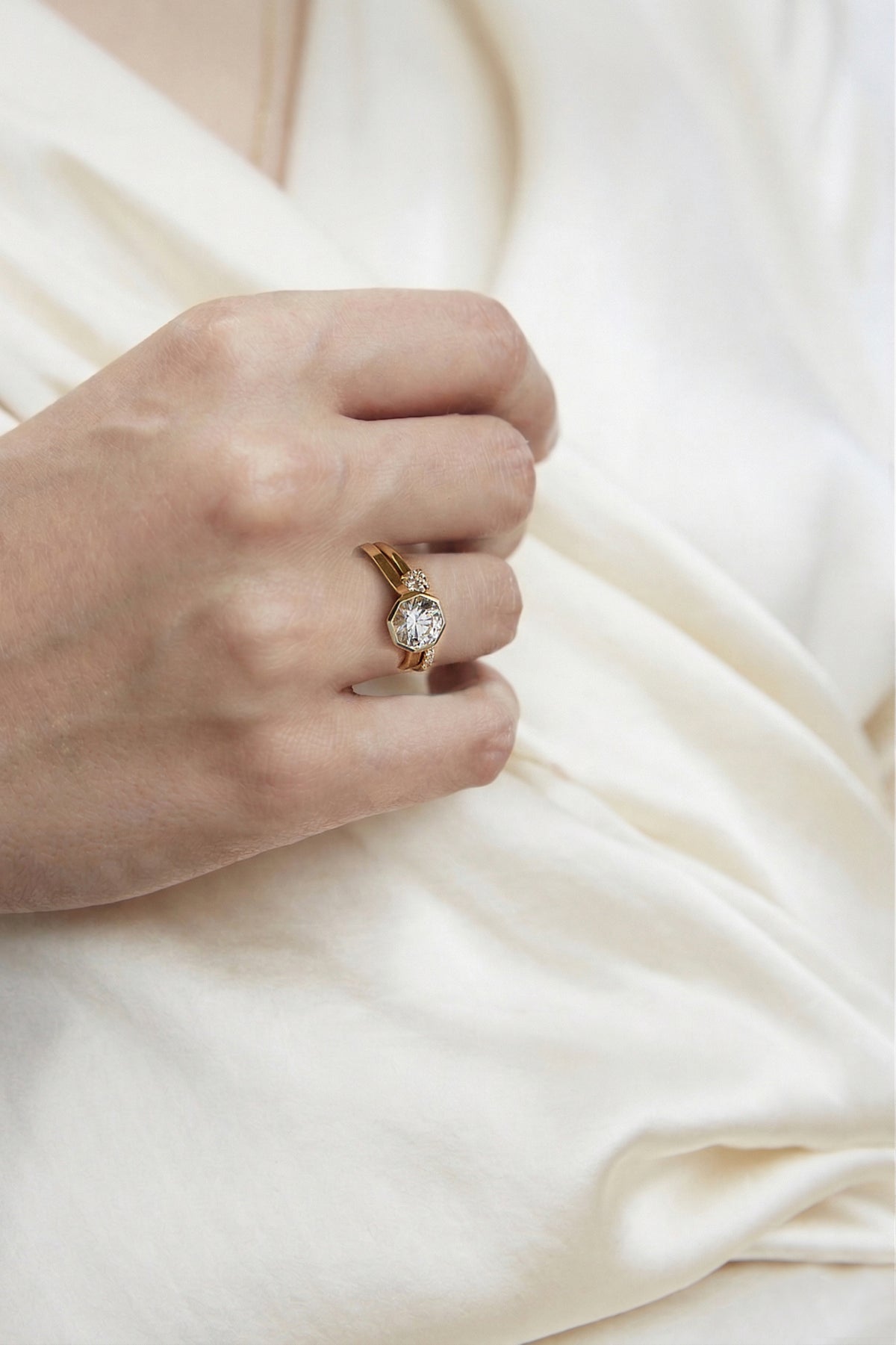 hand wearing a pairing of a 18k yellow gold architectural engagement ring with a round cut diamond in a thick, octagon bezel setting and a open band with a round cut diamond.