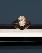 18k yellow gold engagement ring featuring an oval cut diamond in a sculpted octagonal bezel frame against a dark blue background, propped on a reflective surface.