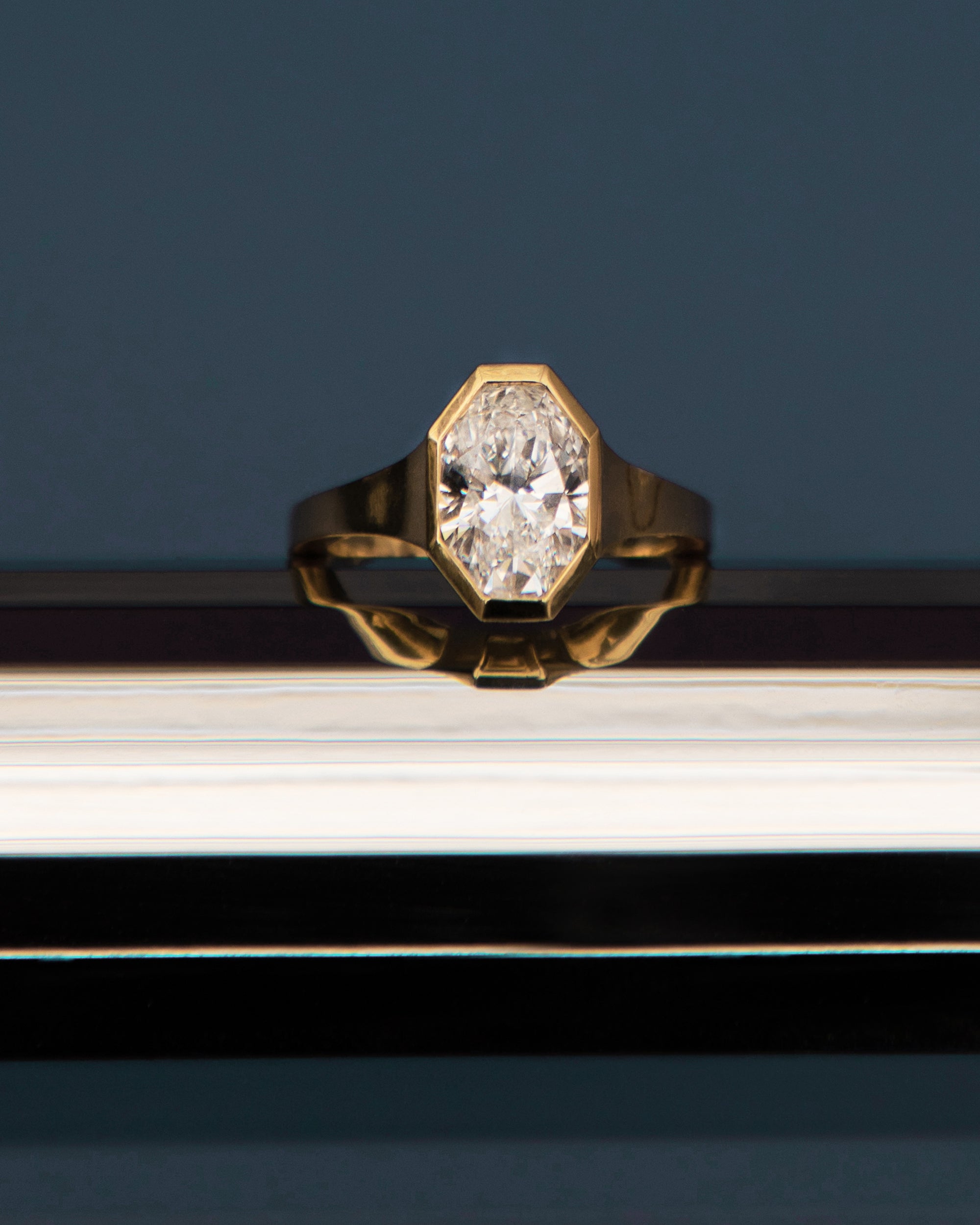 18k yellow gold engagement ring featuring an oval cut diamond in a sculpted octagonal bezel frame against a dark blue background, propped on a reflective surface.