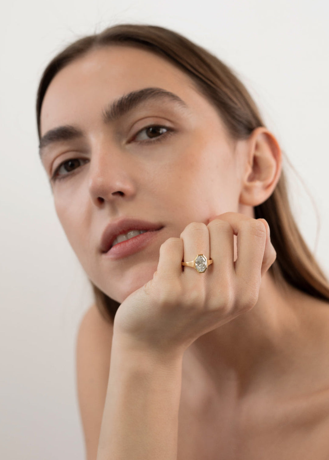 model wearing 18k yellow gold engagement ring featuring an oval cut diamond in a sculpted octagonal bezel frame on her ring finger.