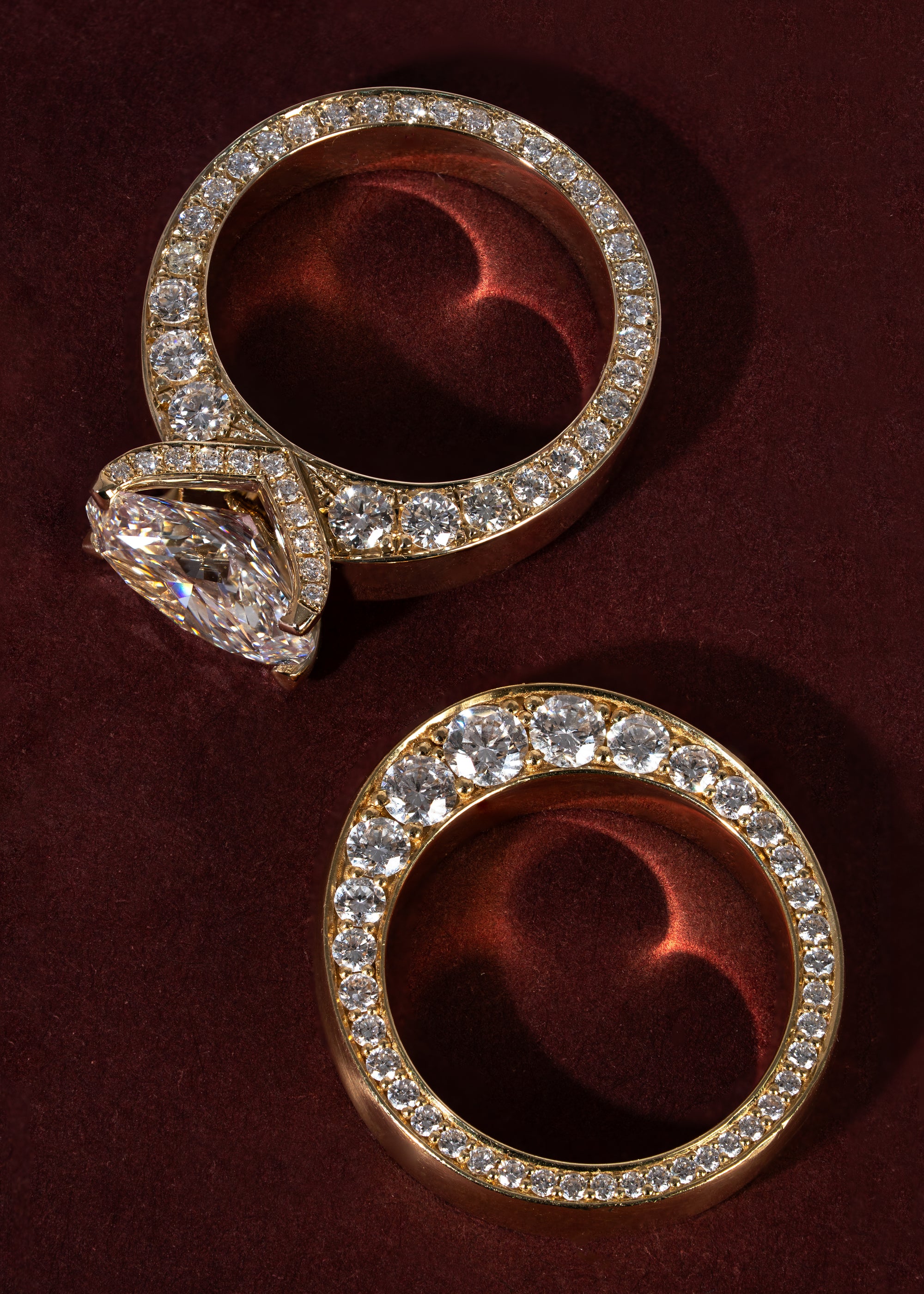 Two thick wide, gold bands with tapering diamonds on the sides of the bands with one of the rings with a oval cut center stone with a V-shaped pavé shank, against a wine colored background.