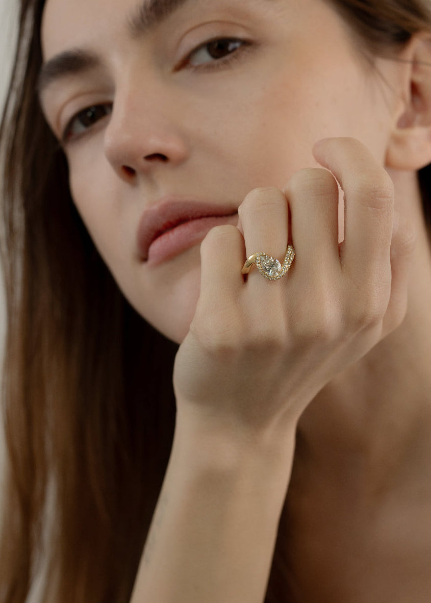 model wearing  18k yellow gold ring featuring a pear-cut diamond in a curved, wave-like setting with pavé diamonds around the center stone