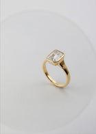 a 18k yellow gold ring with a emerald cut diamond center stone against a light grey background