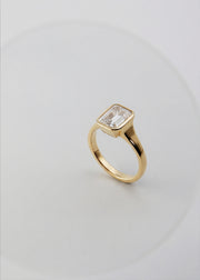 a 18k yellow gold ring with a emerald cut diamond center stone against a light grey background