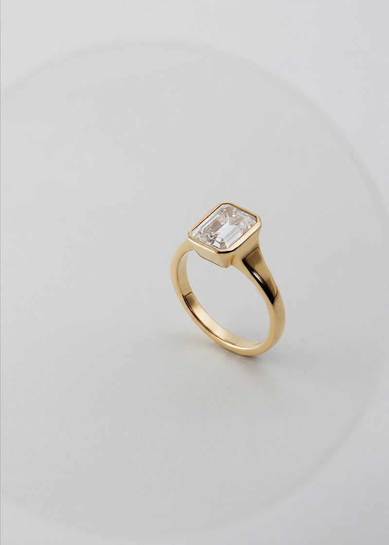 a 18k yellow gold ring with a emerald cut diamond center stone against a light grey background