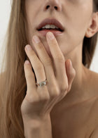 Close-up of a woman's hand with a 18k white gold ring, with a east west emerald cut diamond as the center stone, on her finger, touching her face.