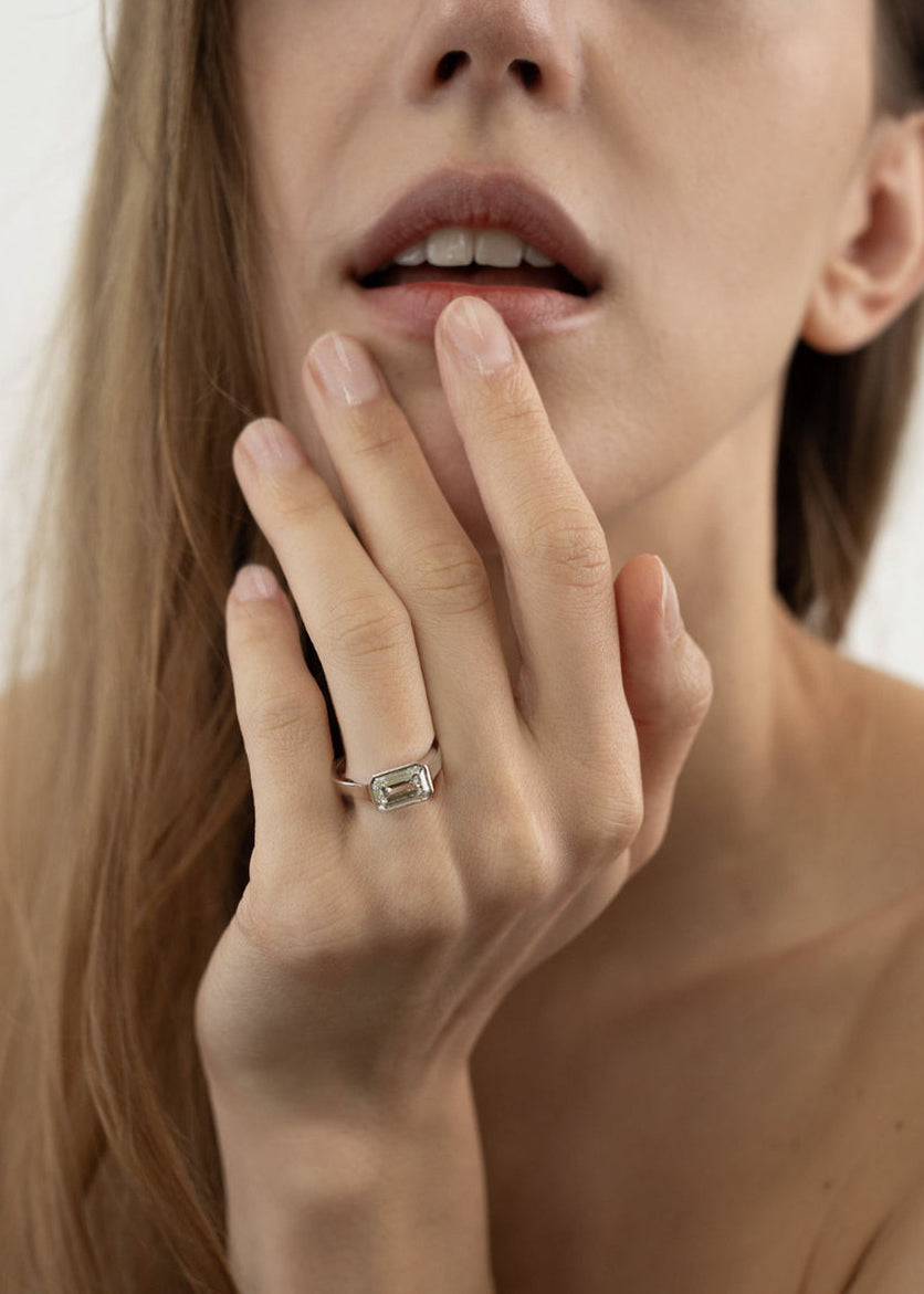 Close-up of a woman's hand with a 18k white gold ring, with a east west emerald cut diamond as the center stone, on her finger, touching her face.