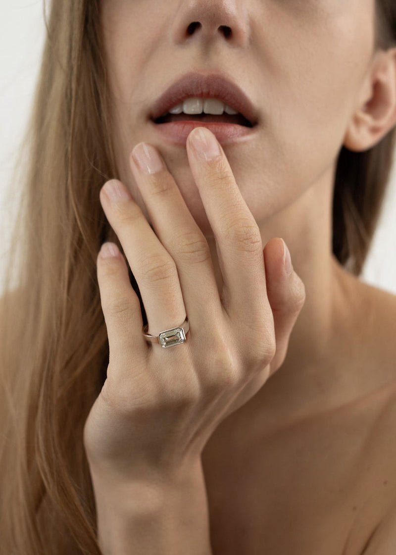 Close-up of a woman's hand with a 18k white gold ring, with a east west emerald cut diamond as the center stone, on her finger, touching her face.
