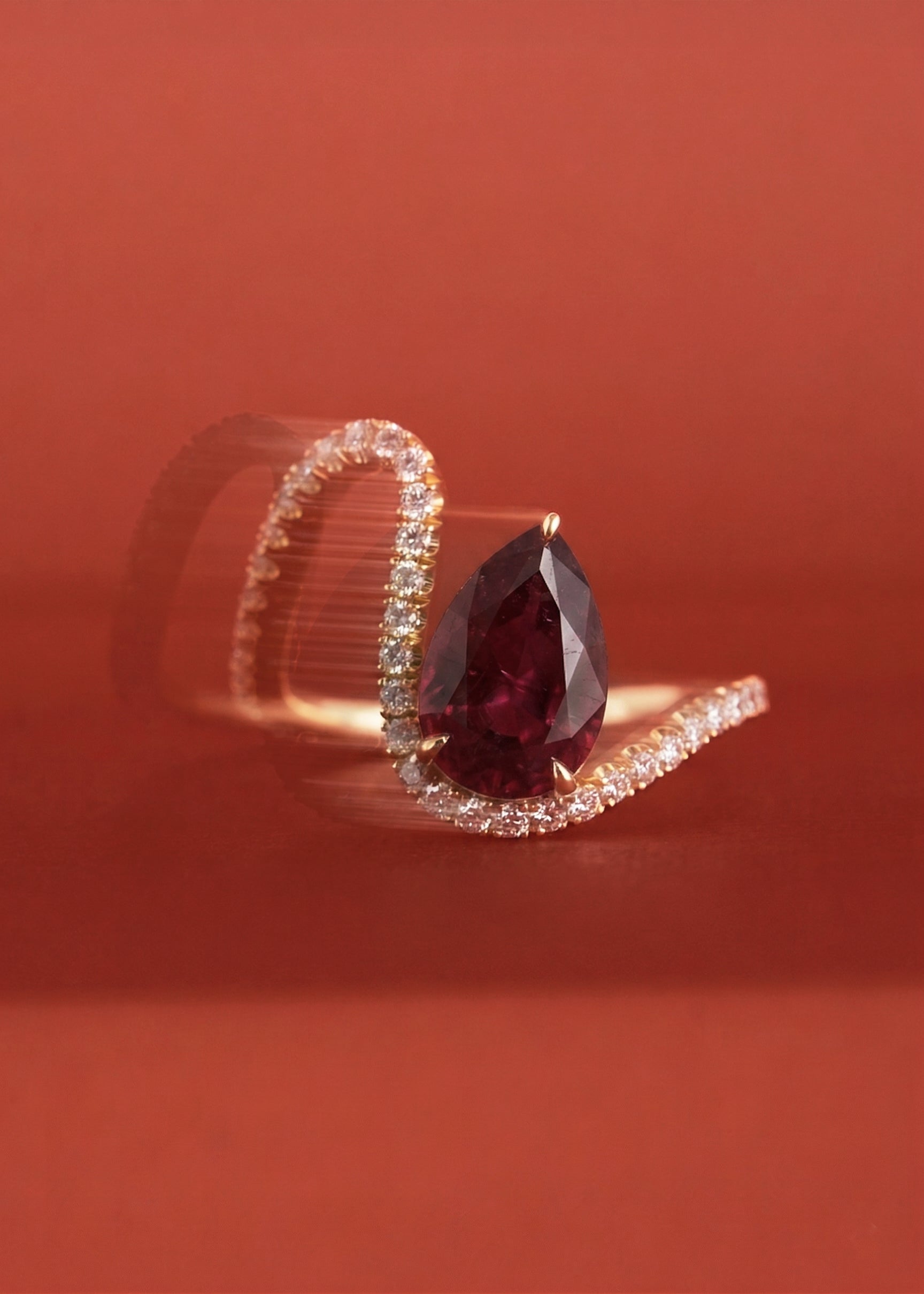 18k yellow gold trace band with pavé diamonds and a floating pear cut ruby slightly tilted- against a pale red background in motion blur
