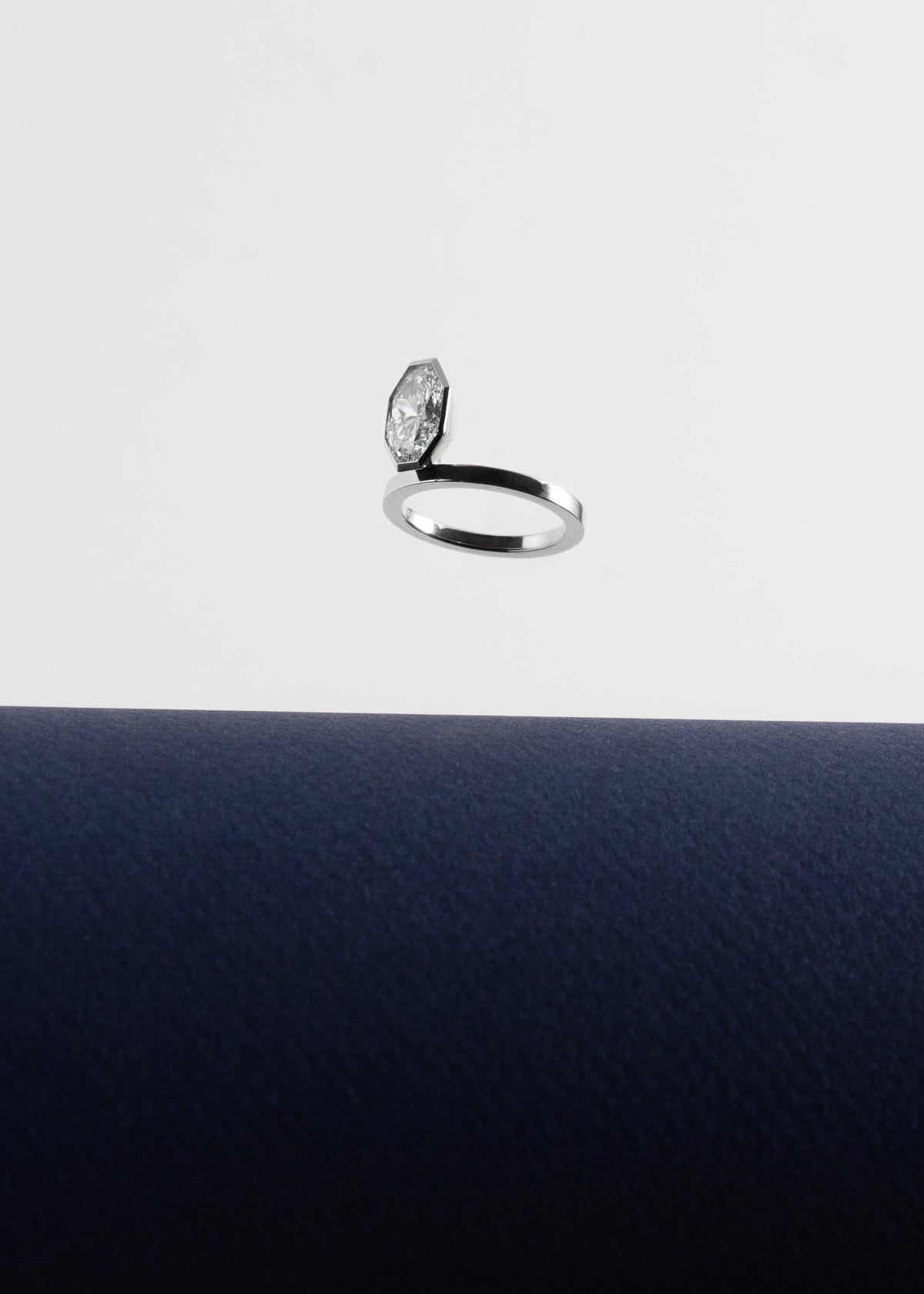 18k white gold engagement ring featuring an elevated floating oval cut diamond in a sculpted octagon bezel setting floating above a dark blue surface