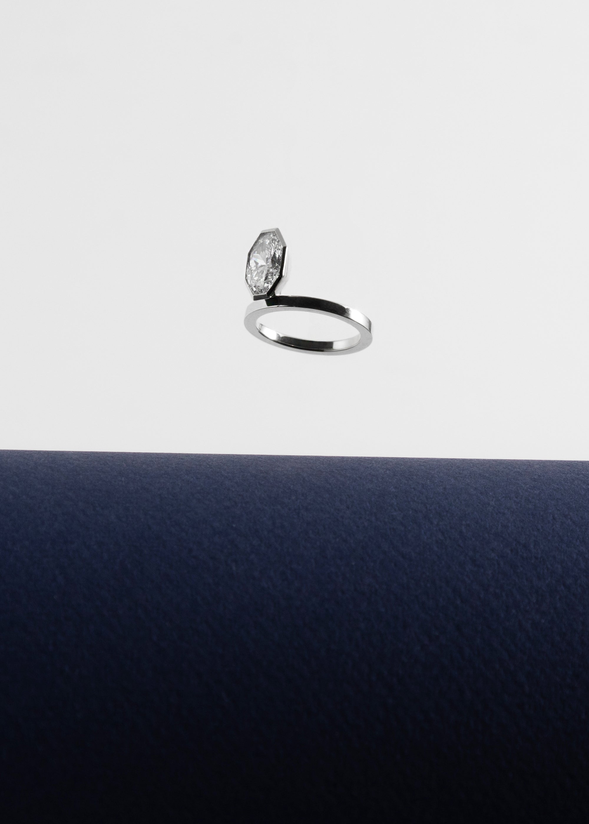 18k white gold engagement ring featuring an elevated floating oval cut diamond in a sculpted octagon bezel setting floating above a dark blue surface