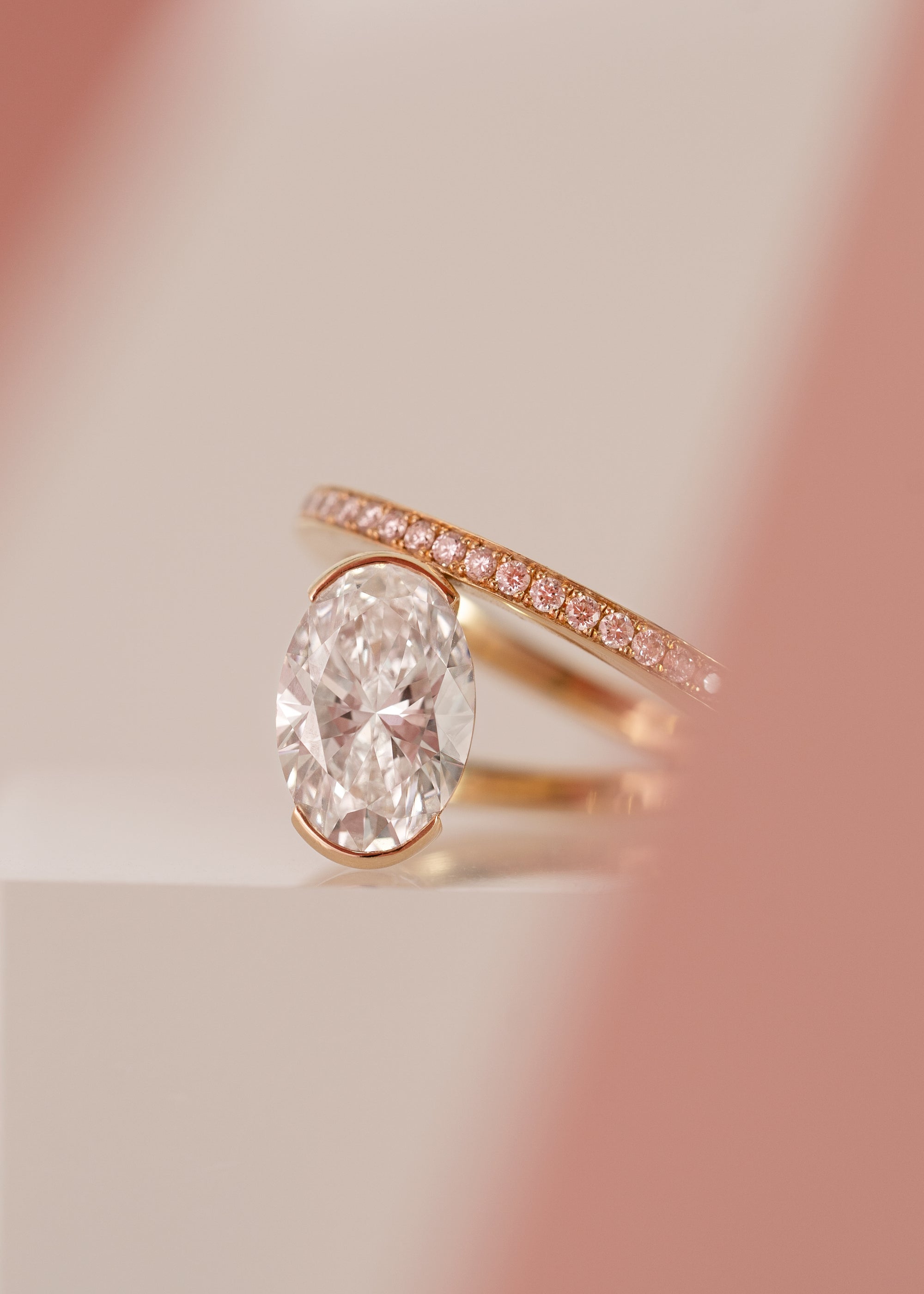 front view- 18k yellow gold ring showcasing a fluid line of faint pink micro-pavé diamonds that winds continuously around the finger- the design securely cradles a floating, bezel-nested faint pink oval diamond along a floating bypass band-KATKIM-engagement ring-fine jewelry