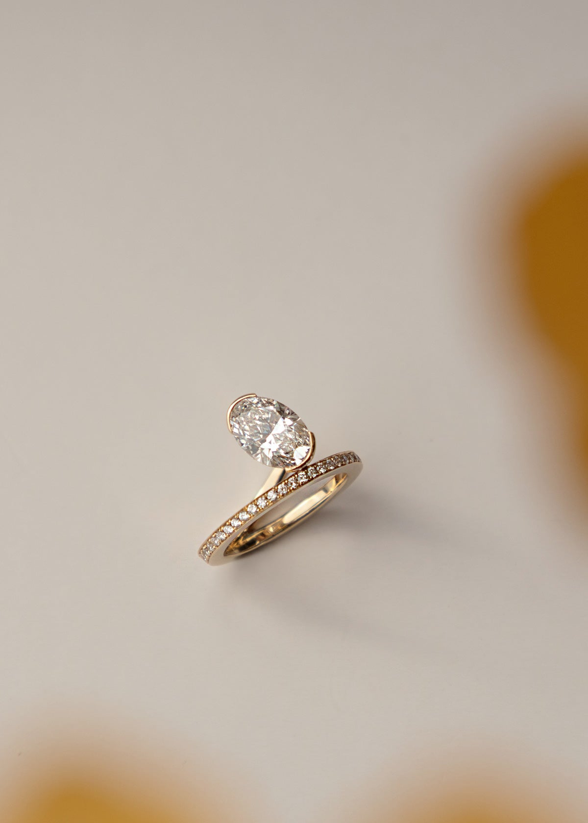 18k yellow gold ring showcasing a fluid line of micro-pavé diamonds that winds continuously around the finger- the design securely cradles a floating, bezel-nested oval diamond along a floating bypass band-aerial view against a light beight background-KATKIM-engagement ring-fine jewelry