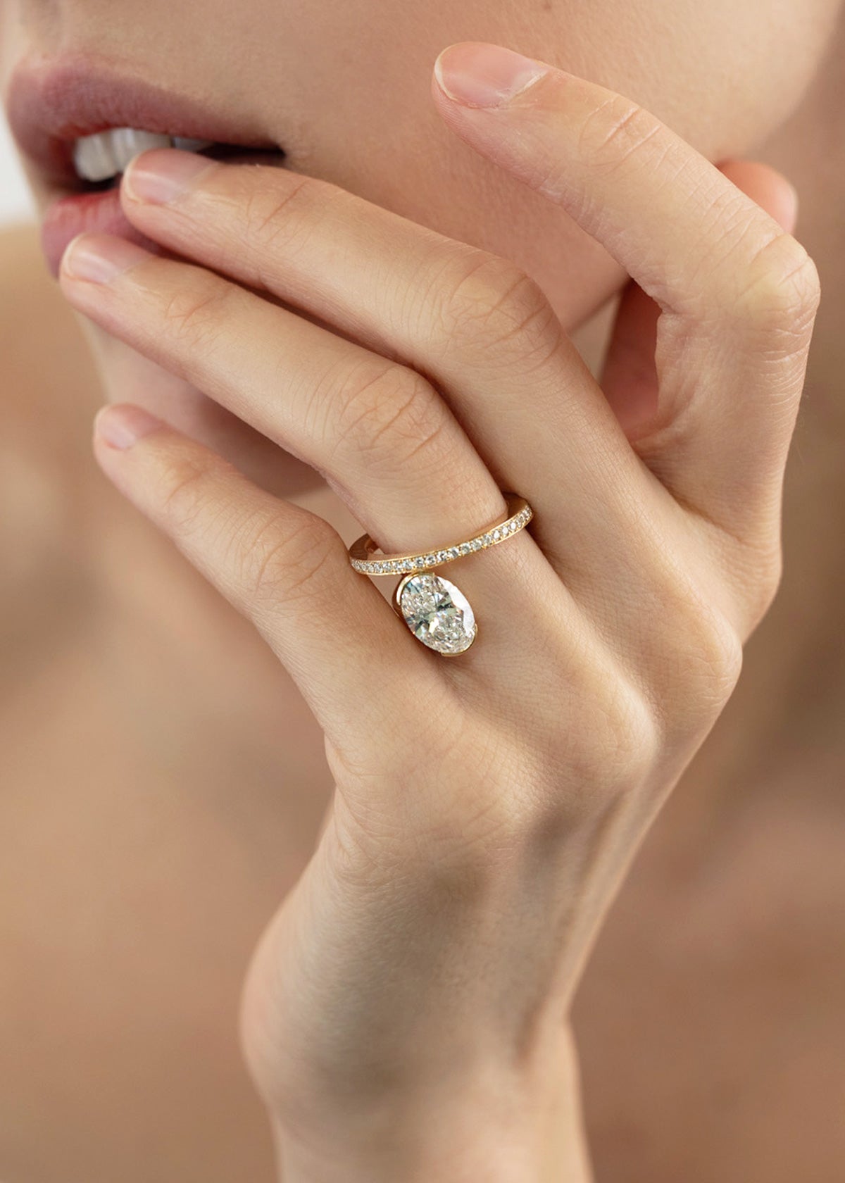 model wearing a 18k yellow gold ring showcasing a fluid line of micro-pavé diamonds that winds continuously around the finger- the design securely cradles a floating, bezel-nested oval diamond along a floating bypass band-ring is worn on the ring finger-hand is nestled slightly aginst the model's chin-KATKIM-engagement ring-fine jewelry