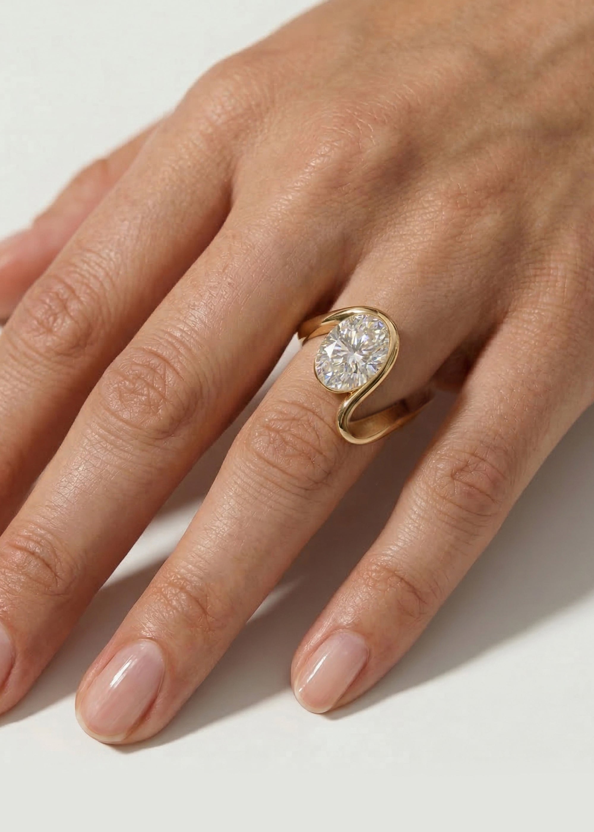 hand wearing a 18k yellow gold ring with a wide wavy band resembling a ribbon nestling an oval cut diamond