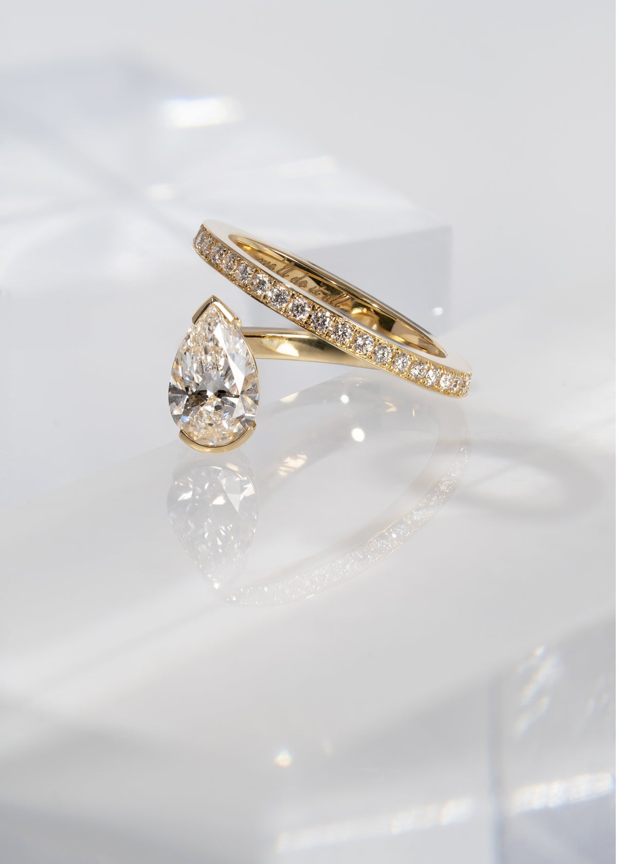 18k yellow gold engagement ring featuring a floating pear cut diamond on an architectural tilted micropavé band against a reflective surface.