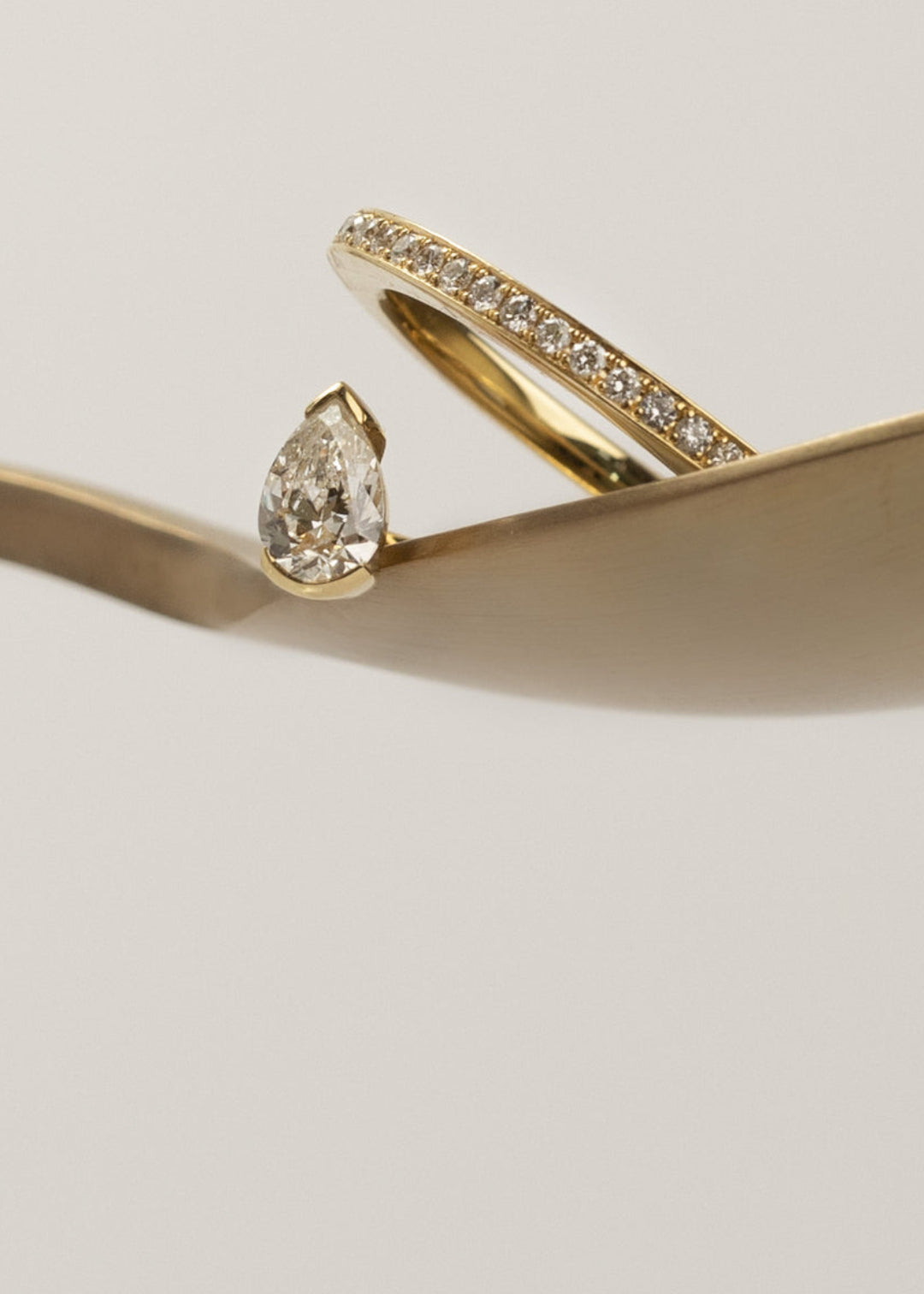 18k yellow gold engagement ring featuring a floating pear cut diamond on an architectural tilted micropavé band held on a curved brown surface.