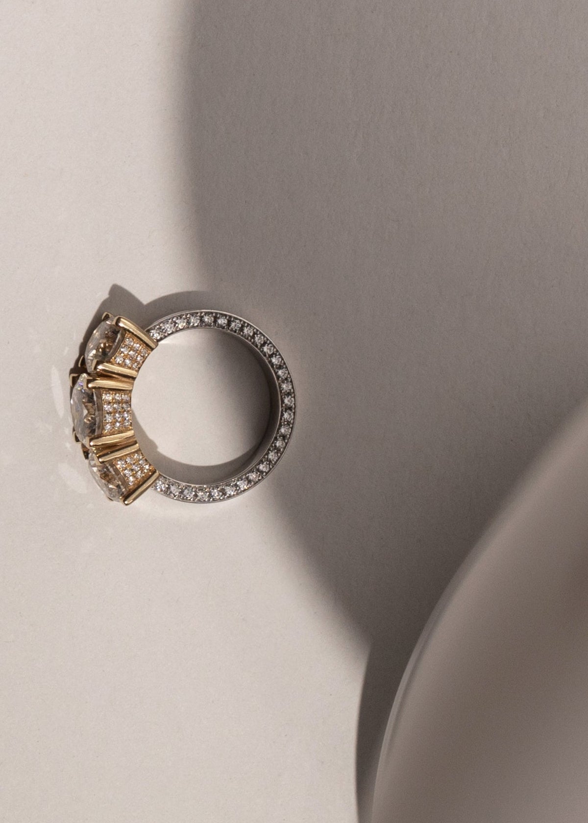 Close-up of a mixed metal ring a pavé diamonds on the band with the three round diamonds with halo set pave diamonds around with intricate design on a neutral background