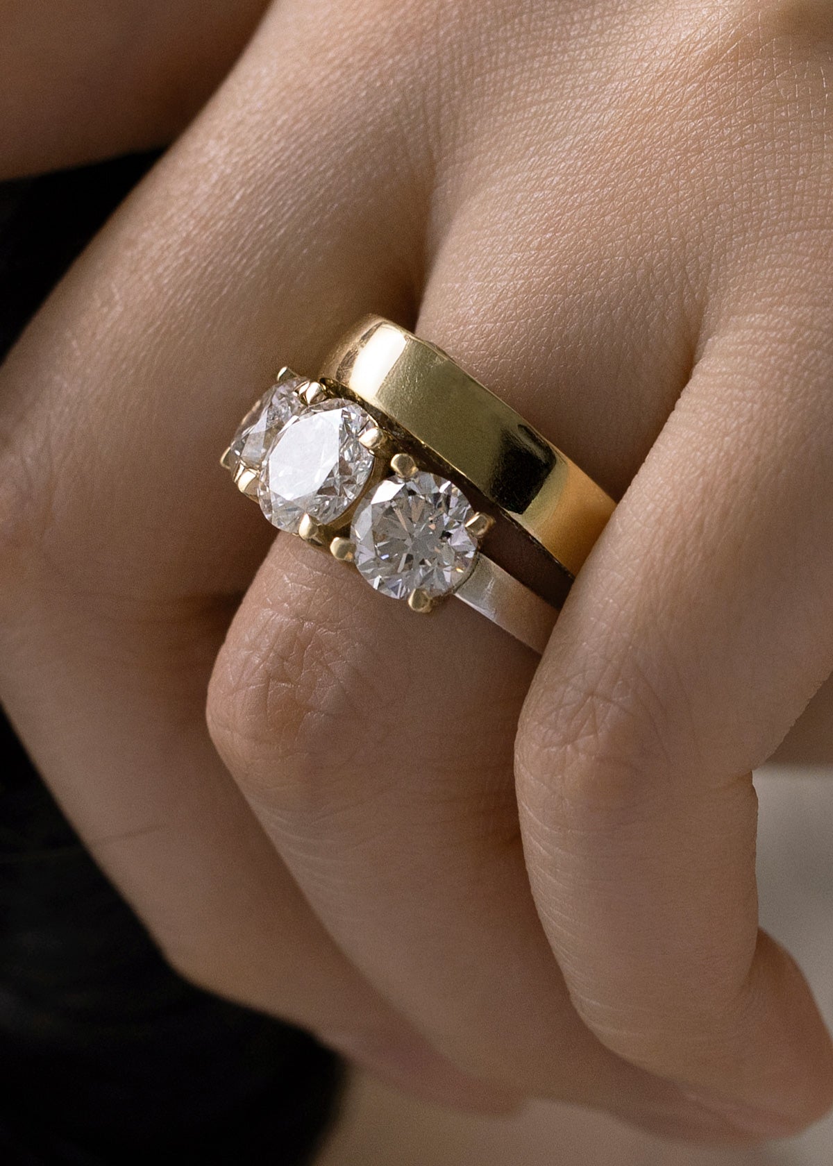 close-up of a hand wearing two rings, one is a thick gold band and another is thinner white gold band with three round cut diamonds.