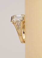 A yellow gold ring featuring a central asscher-cut diamond flanked by two baguette-cut side stones, set horizontally against a light beige background