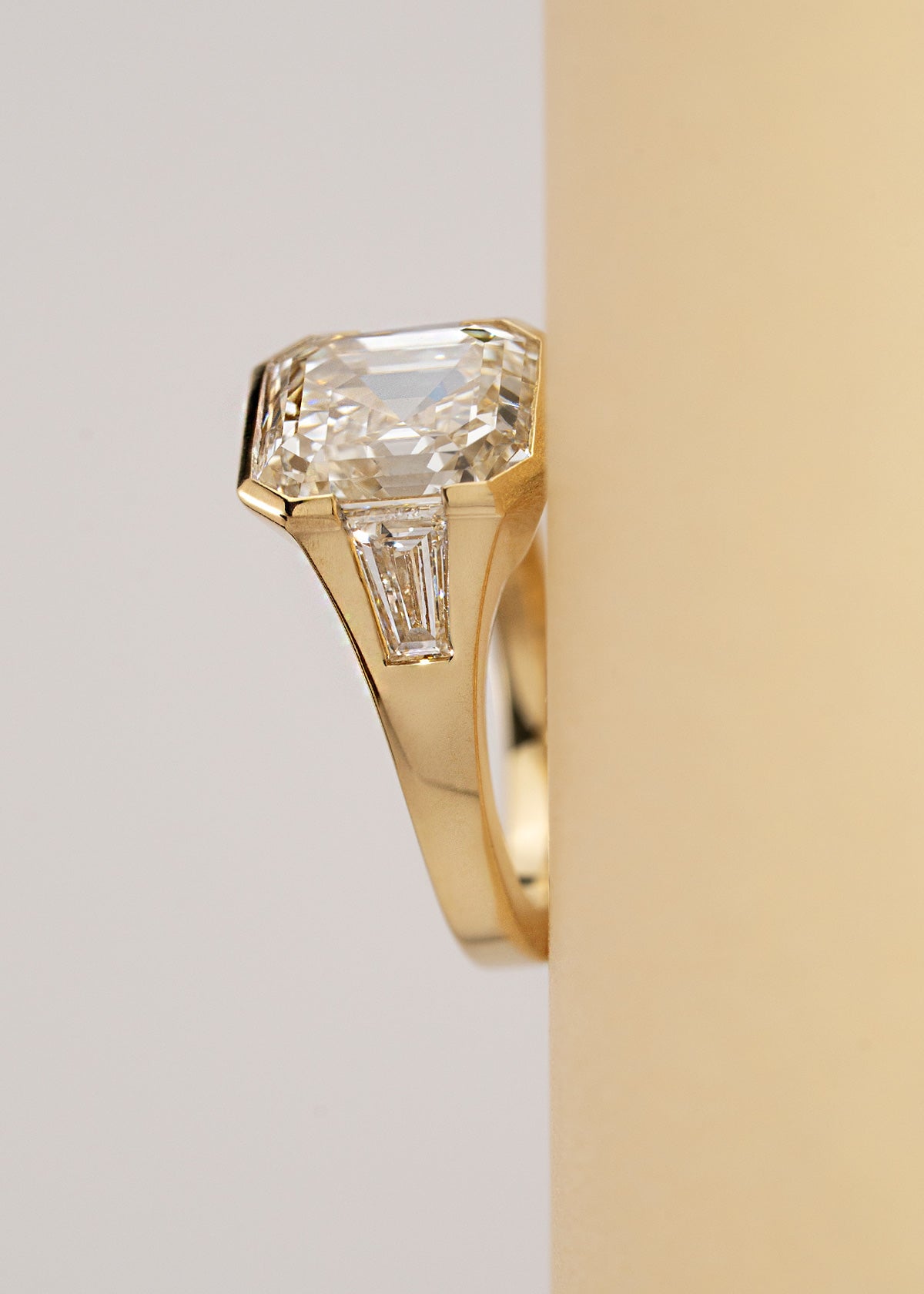 A yellow gold ring featuring a central asscher-cut diamond flanked by two baguette-cut side stones, set horizontally against a light beige background