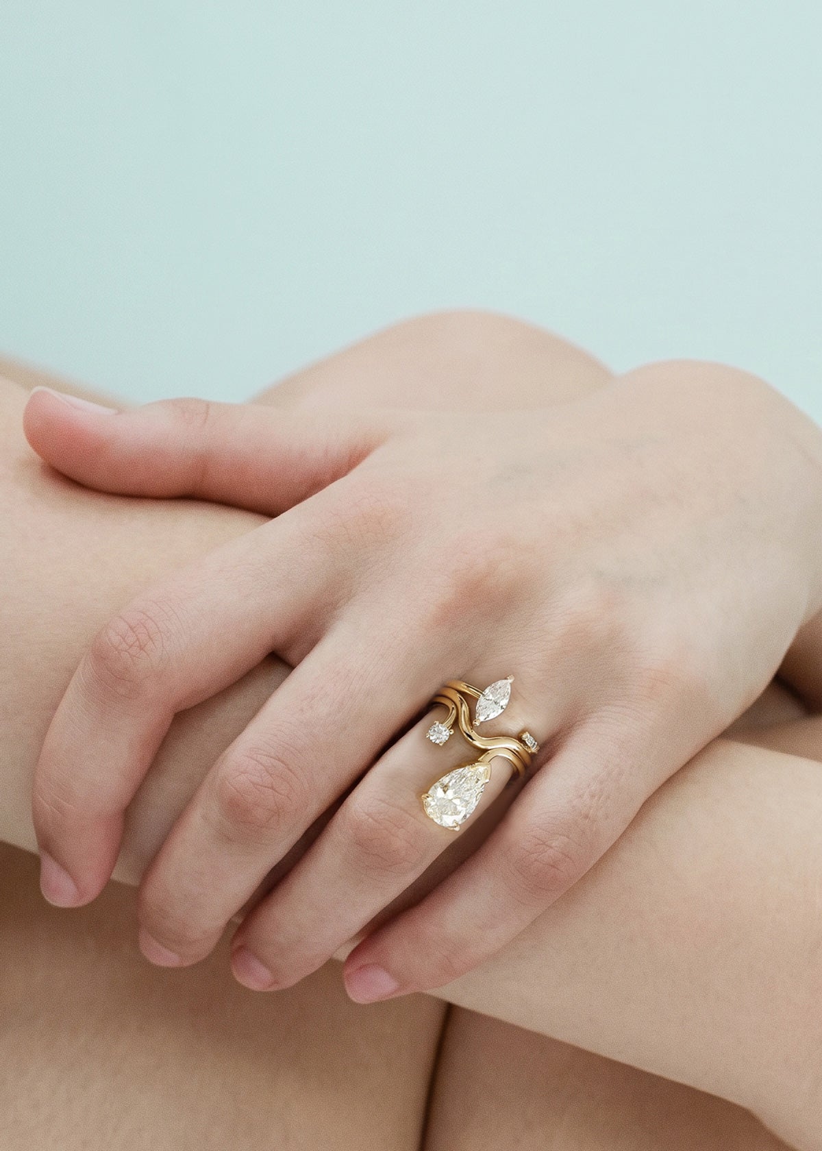 Close-up of a hand wearing three 18k yellow rings–one with a large pear-cut diamond drop and a smaller round diamond accent on a curved band, curved gold band, open band with a marquise cut diamond.