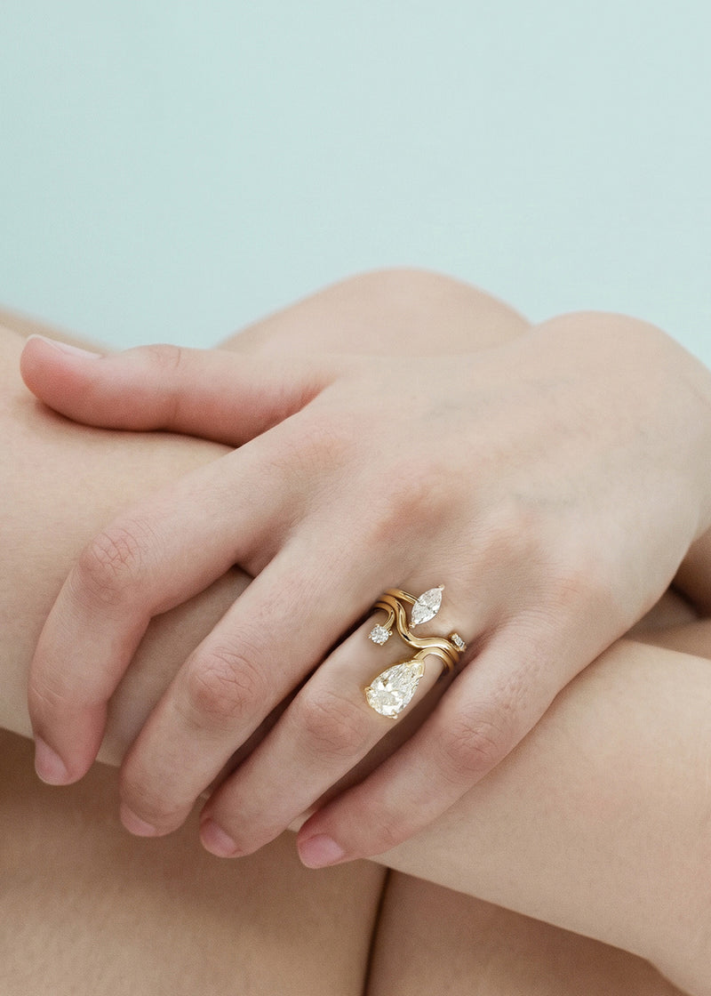 Close-up of a hand wearing three 18k yellow rings–one with a large pear-cut diamond drop and a smaller round diamond accent on a curved band, curved gold band, open band with a marquise cut diamond.
