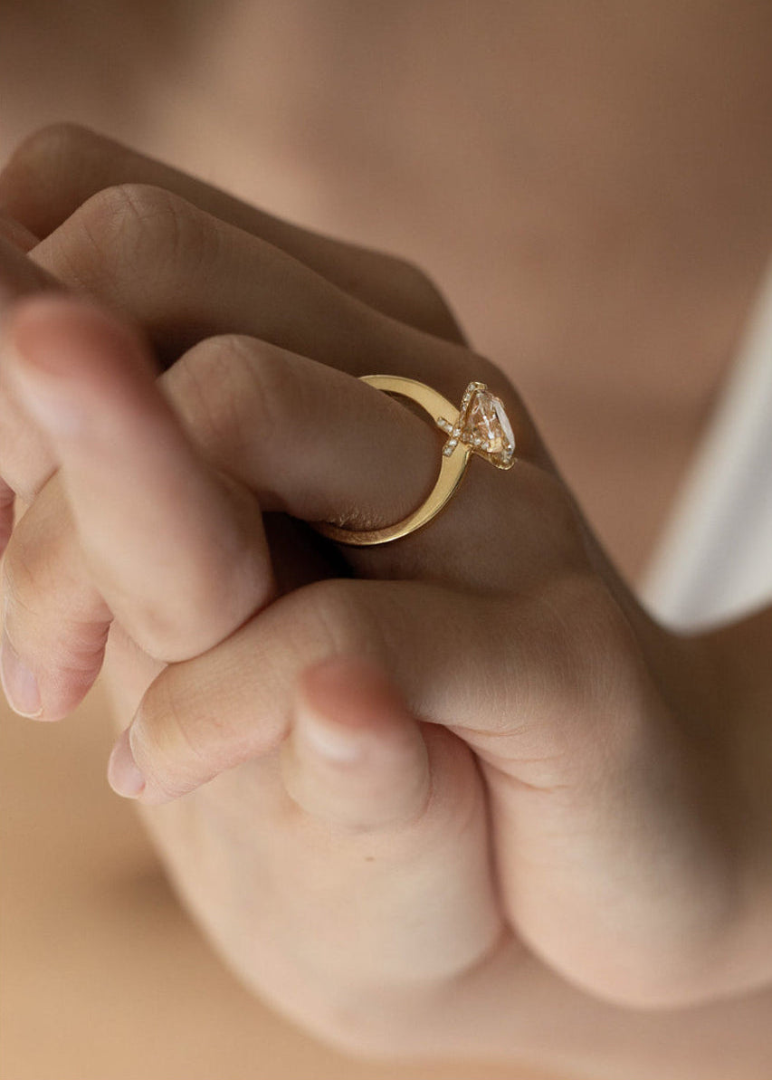 Close-up of a hand wearing a 18k yellow gold solitaire engagement ring featuring a oval cut diamond  accented by small pavé diamonds on the x-shaped shank.