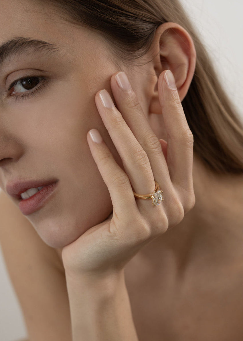 Close-up of a woman's hand wearing a 18k yellow gold solitaire engagement ring featuring a oval cut diamond  accented by small pavé diamonds on the x-shaped shank.