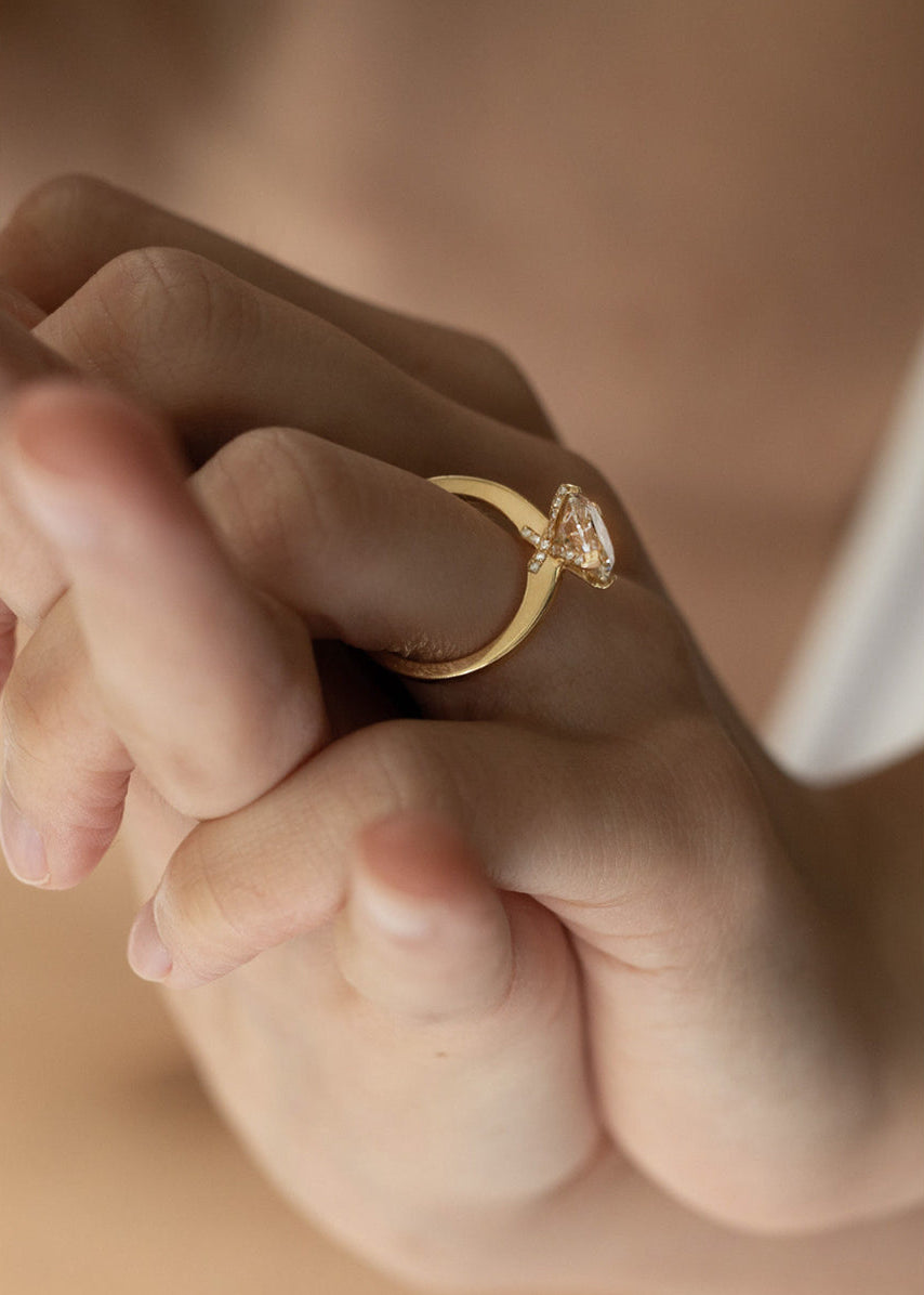 Close-up of a hand wearing a 18k yellow gold solitaire engagement ring featuring a oval cut diamond  accented by small pavé diamonds on the x-shaped shank.