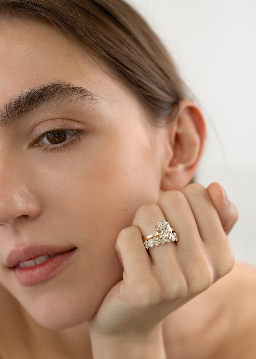Close-up of a hand wearing a 18k yellow gold solitaire engagement ring featuring a oval cut diamond  accented by small pavé diamonds on the x-shaped shank paired with a band of an eternity of oval diamonds
