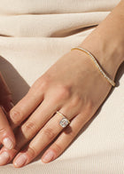 Close-up of a hand wearing a 18k yellow gold engagement ring with a suspended floating asscher cut diamond and half pavé band on a beige fabric background