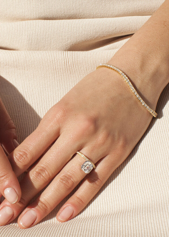 Close-up of a hand wearing a 18k yellow gold engagement ring with a suspended floating asscher cut diamond and half pavé band on a beige fabric background